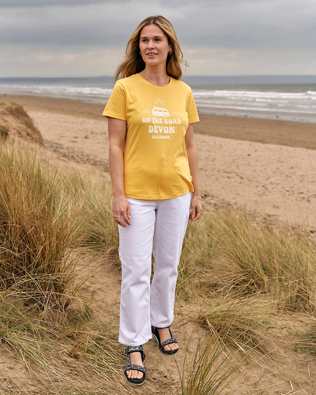 A woman standing on the beach wearing a Saltrock On The Road Devon - Womens Short Sleeve T-Shirt - Yellow.