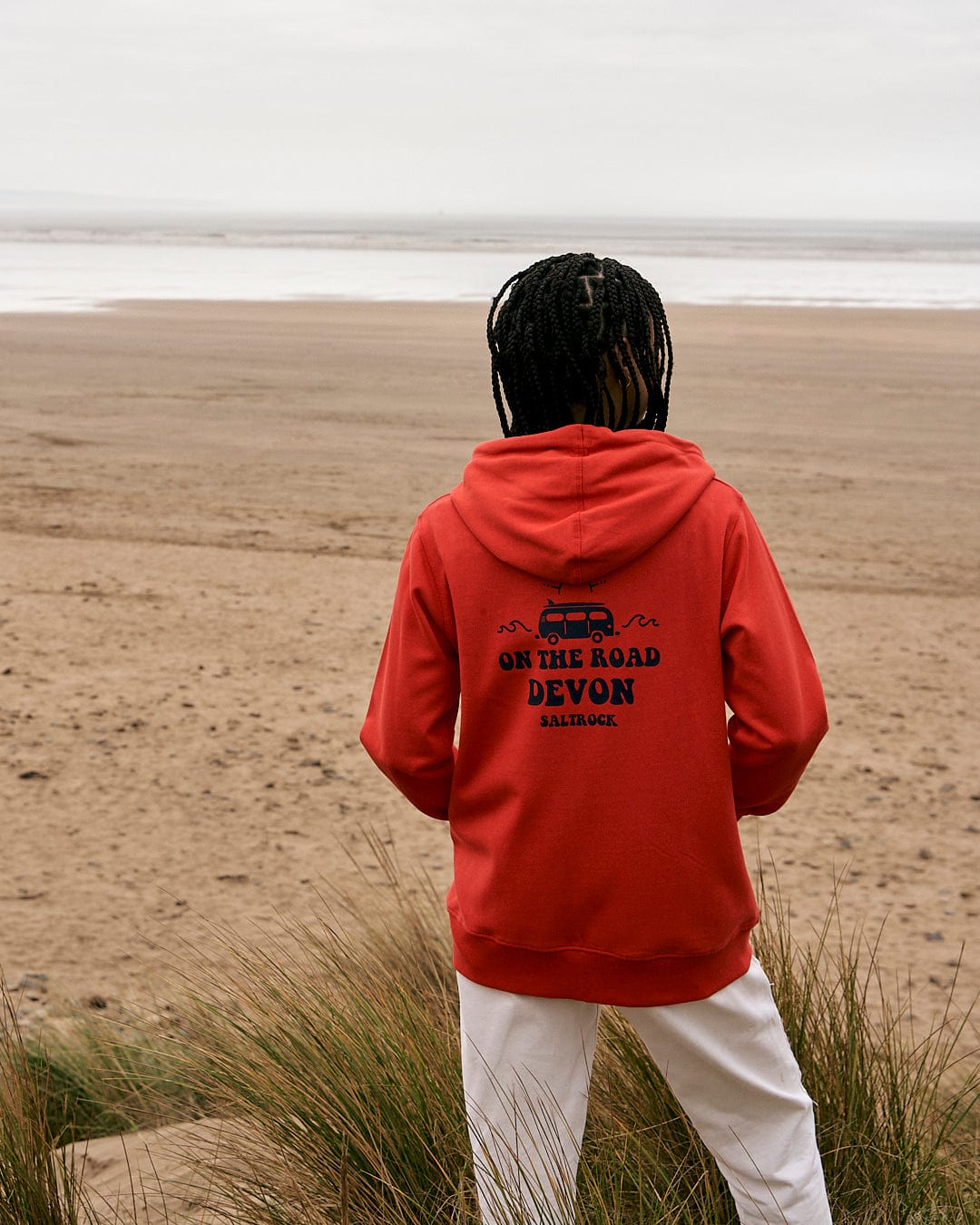 A woman wearing a Saltrock On The Road Devon - Womens Zip Hoodie - Red standing on the beach.