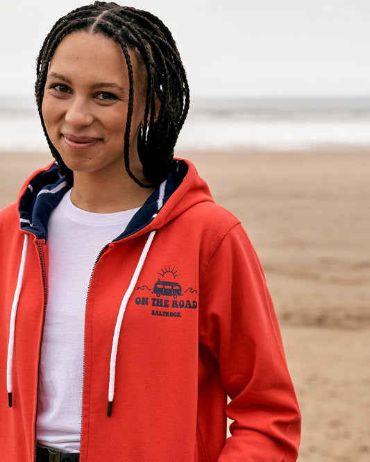 A woman wearing a Saltrock On The Road Devon - Womens Zip Hoodie - Red on the beach.
