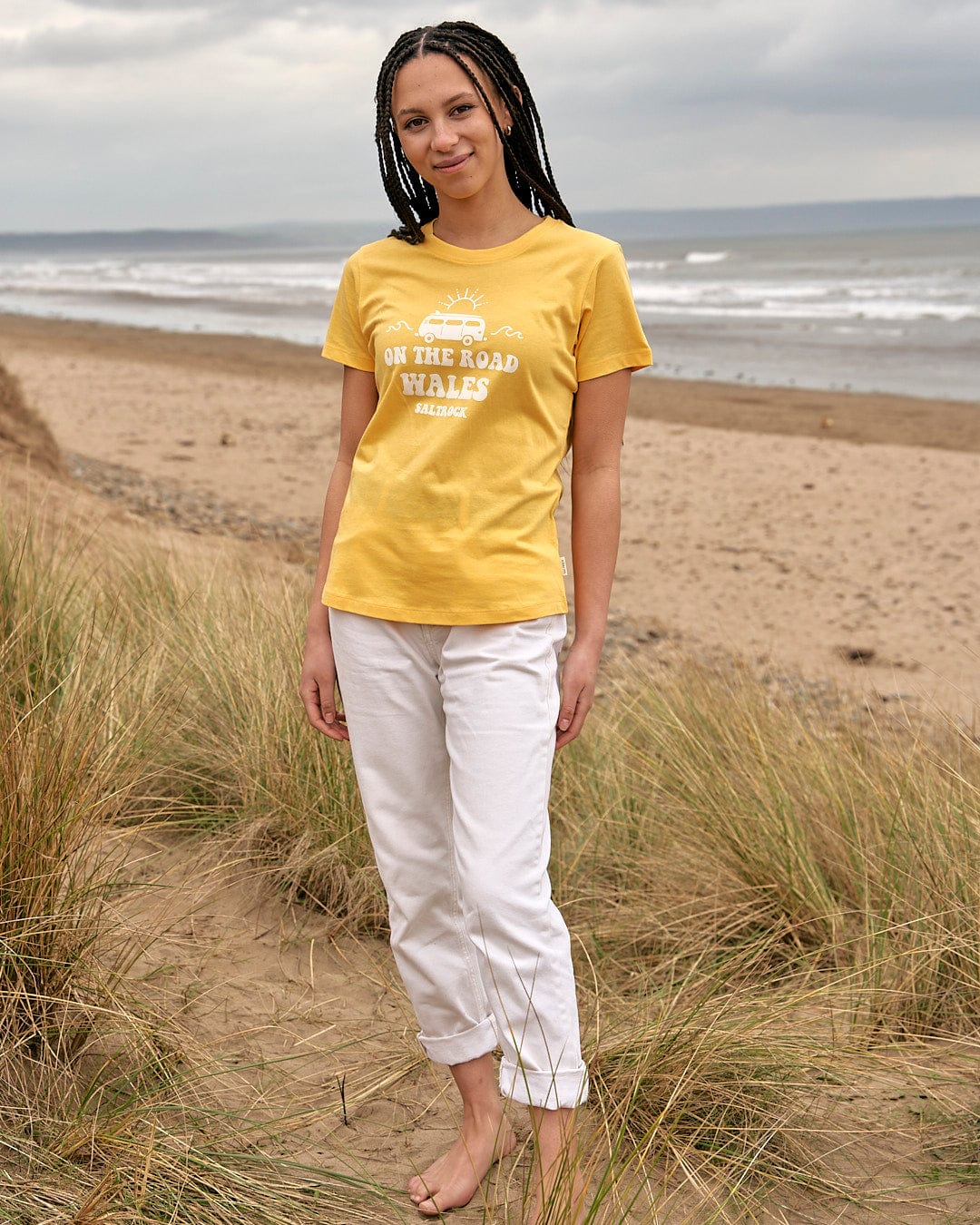A woman wearing a Saltrock On The Road Wales - Womens Short Sleeve T-Shirt - Yellow standing on the beach.