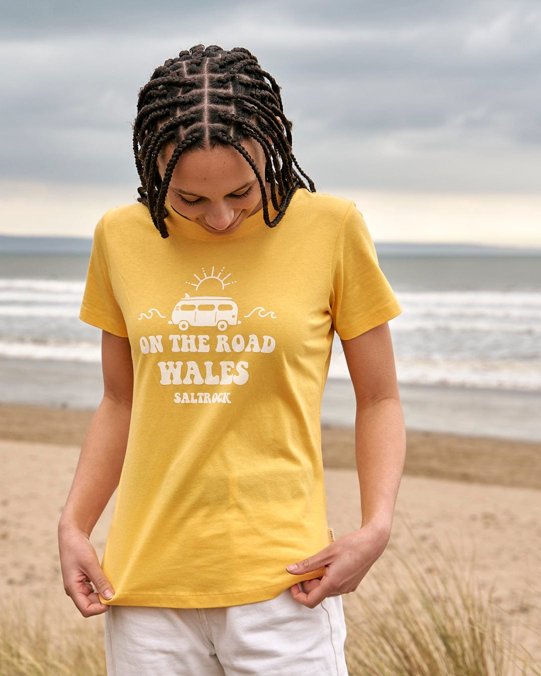 A woman wearing a Saltrock On The Road Wales - Womens Short Sleeve T-Shirt - Yellow standing on the beach.