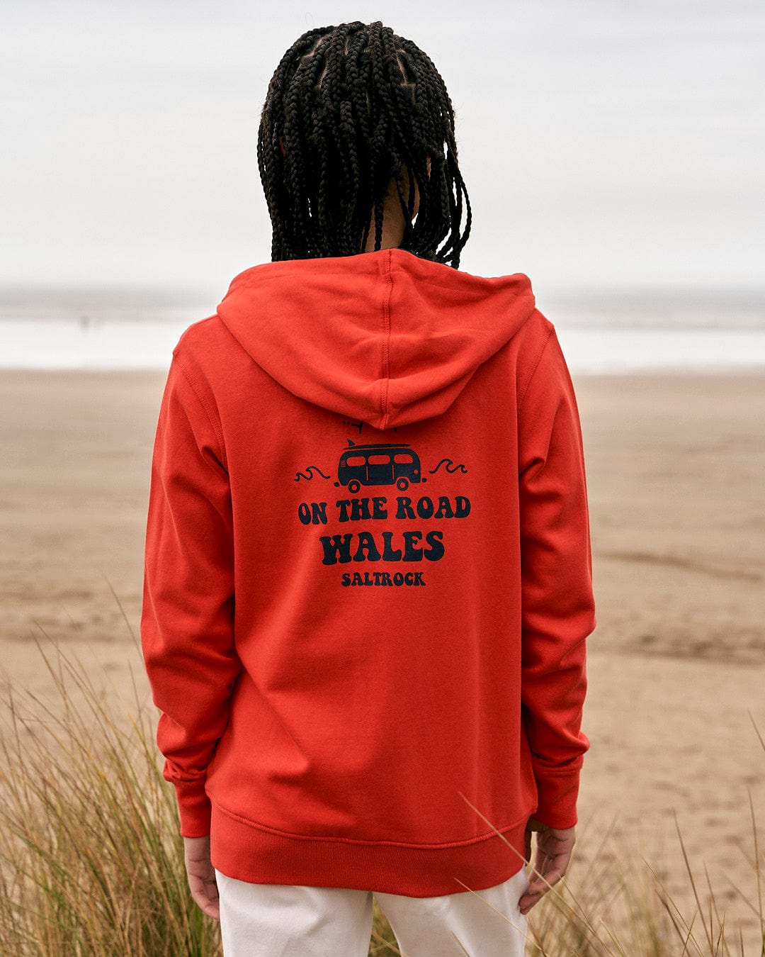 A woman wearing a Saltrock On The Road Wales - Women's Zip Hoodie - Red standing on the beach.