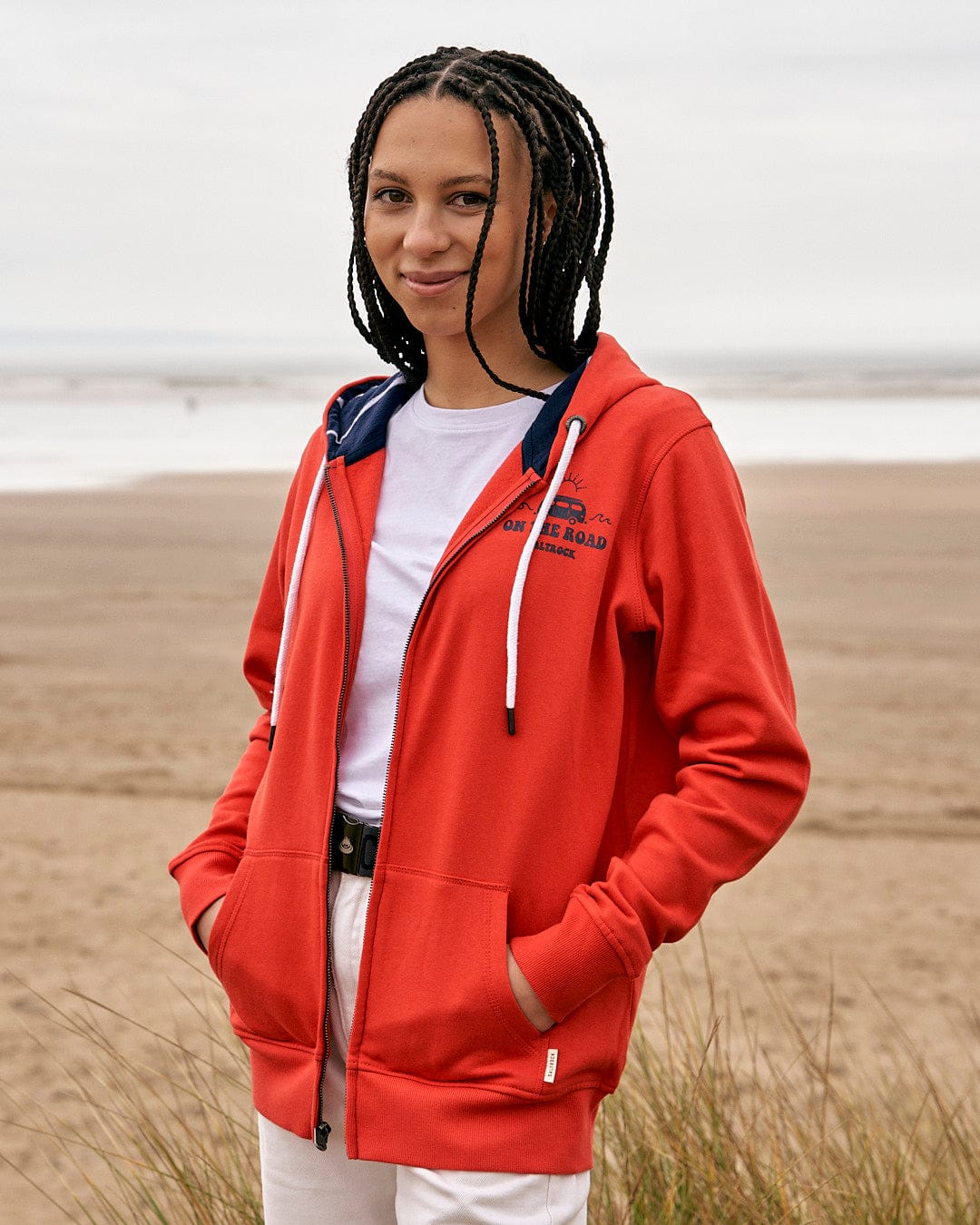 A woman in a Saltrock On The Road Wales - Womes Zip Hoodie - Red standing on a beach.