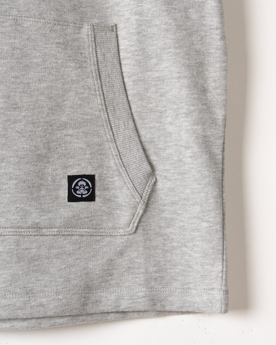 Close-up of a grey melange fabric texture with a focus on the stitched hem and a small black and white Saltrock graphic on the SR Original - Kids Sleeveless Pop Hoodie - Grey clothing label.