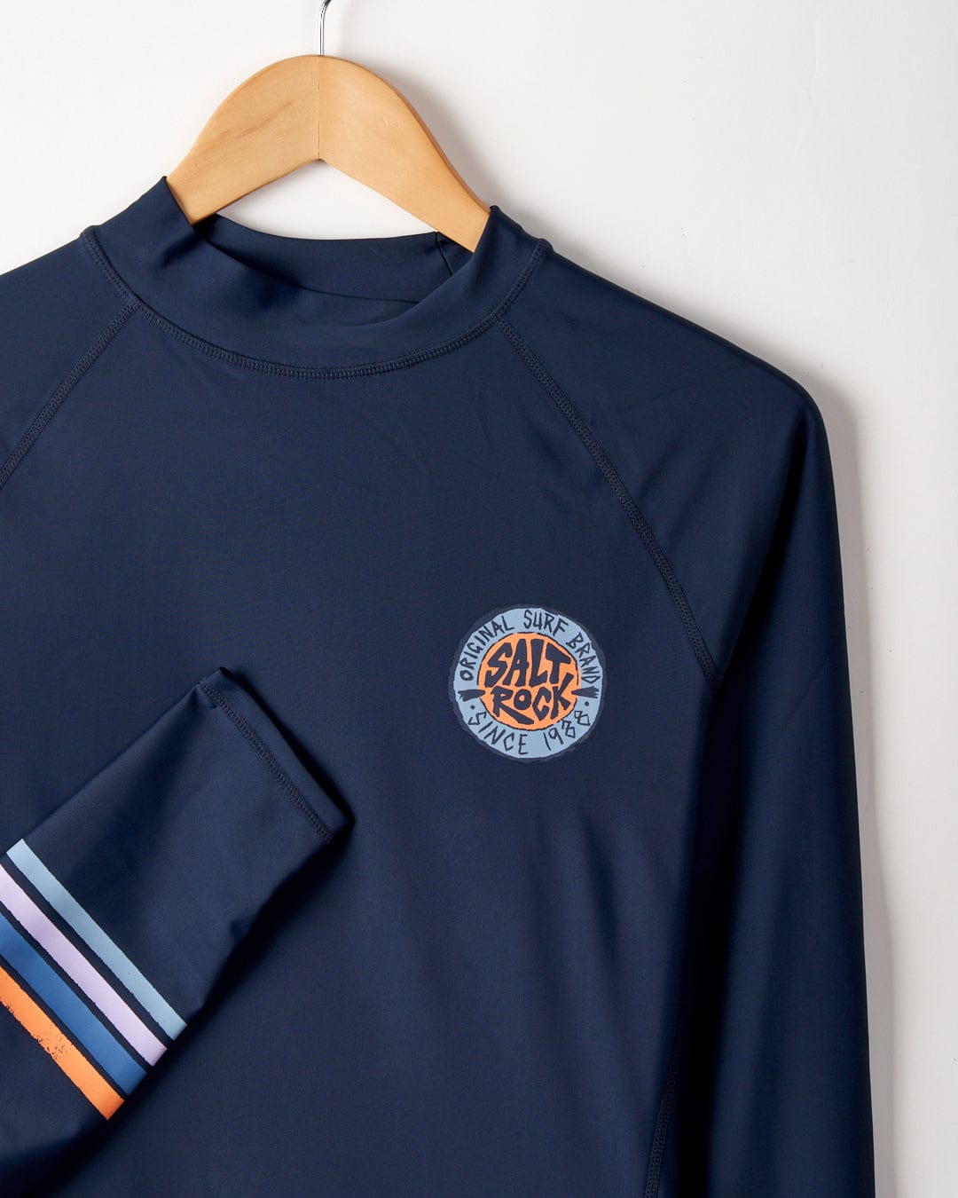 Saltrock's SR Original - Recycled Mens Long Sleeve Rashvest in Navy Blue with UPF 50 protection hanging on a wooden hanger against a white wall, featuring a logo on the chest and striped detail on the sleeve.