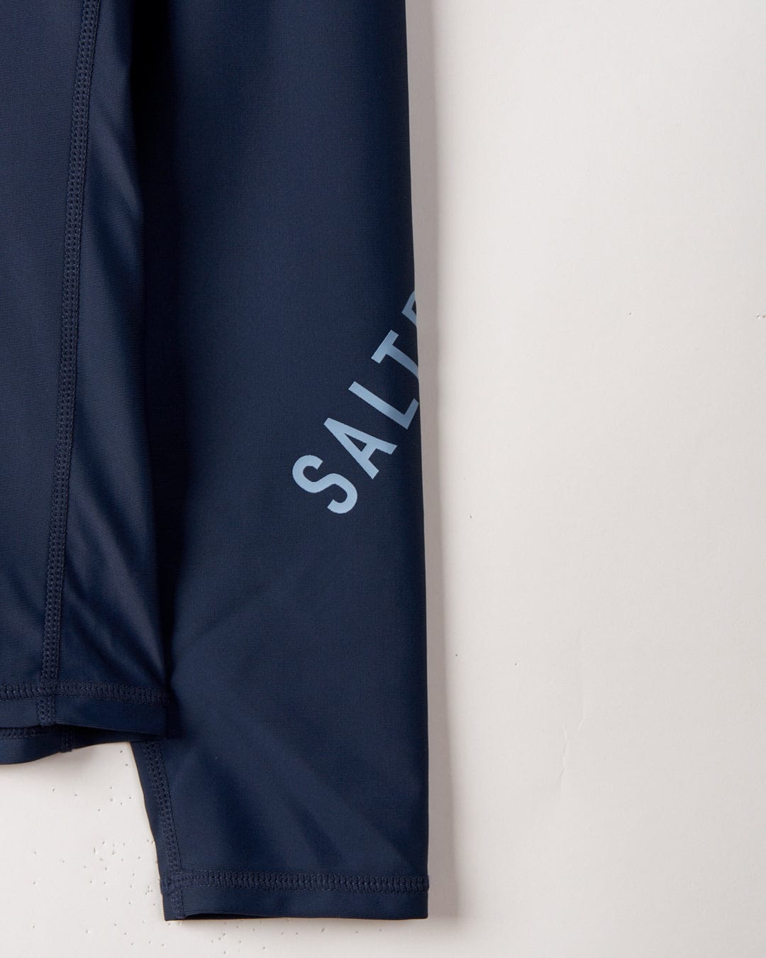 Close-up of SR Original - Recycled Mens Long Sleeve Rashvest - Blue with the word "salt" printed in light gray letters, hanging against a textured white wall.