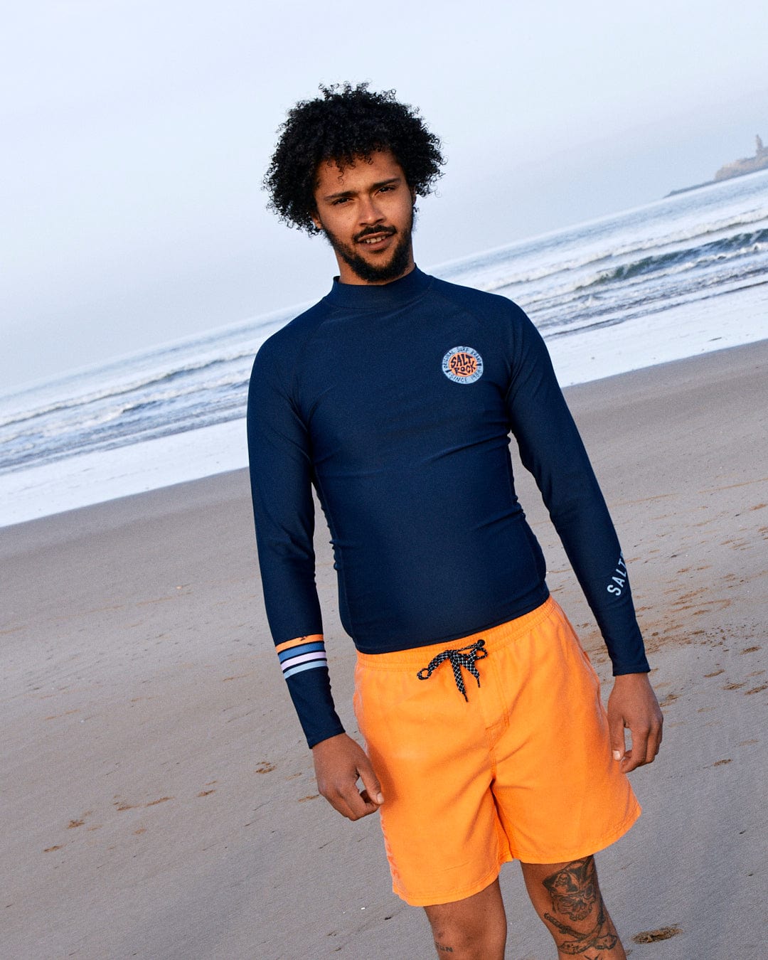 Man in a Saltrock SR Original - Recycled Mens Long Sleeve Rashvest - Blue and orange boardshorts made of Recycled Polyester, standing on a beach with the ocean in the background.