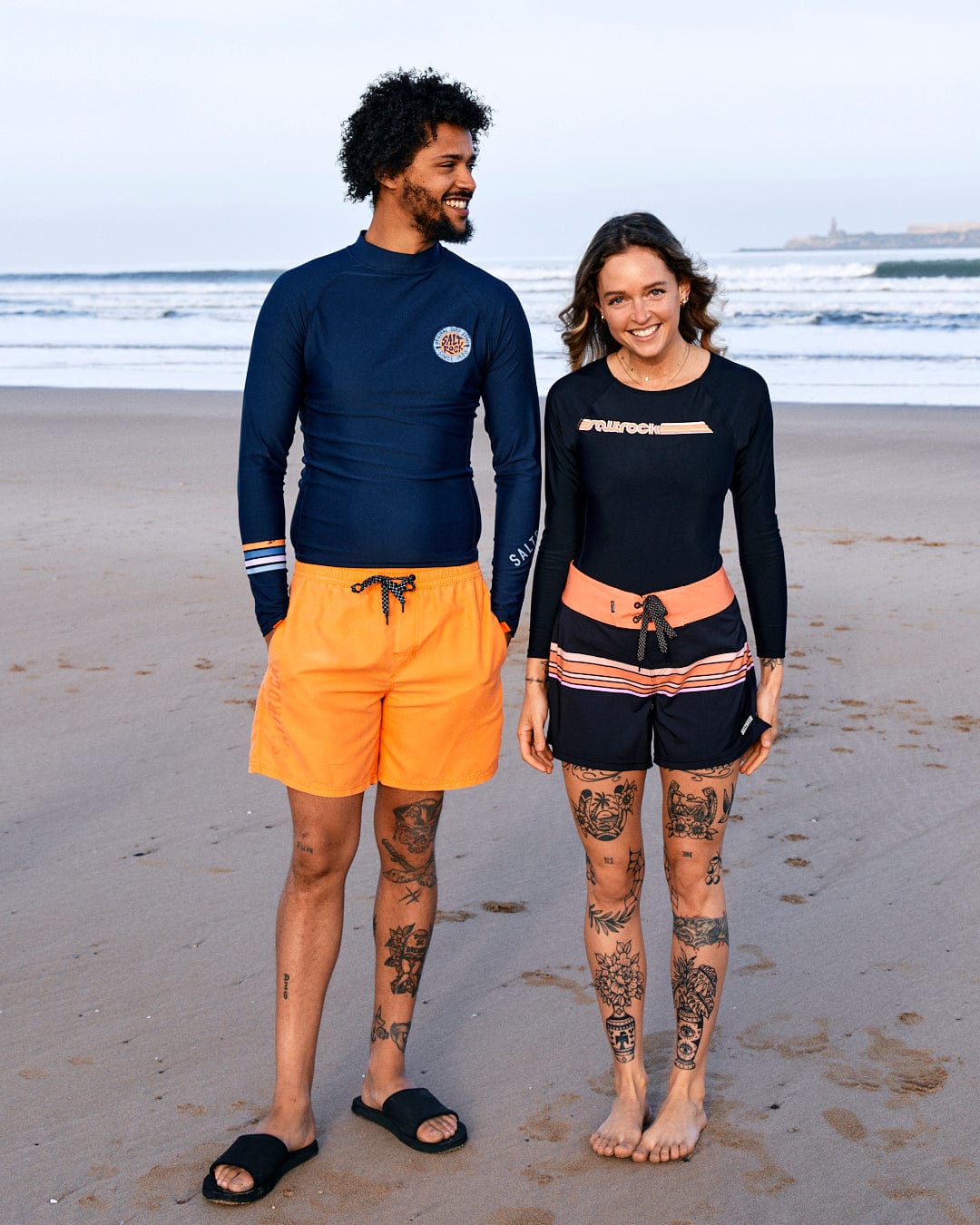 A man and woman smiling on a beach, dressed in Saltrock UPF 50 protection boardshorts and SR Original - Recycled Mens Long Sleeve Rashvest - Blue, with visible tattoos on their legs.