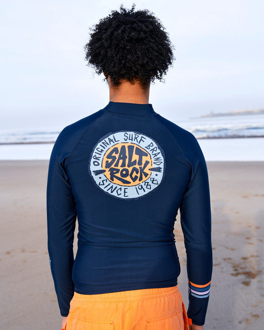 Person wearing a Saltrock SR Original - Recycled Mens Long Sleeve Rashvest in Blue with UPF 50 protection and "salt rock original surf park since 1948" logo on the back, standing on a sandy beach.
