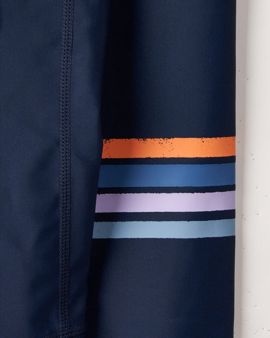 Close-up of a Saltrock SR Original - Recycled Mens Long Sleeve Rashvest - Blue with horizontal stripes in orange, blue, and purple, designed for UPF 50 protection, hanging next to a white column.