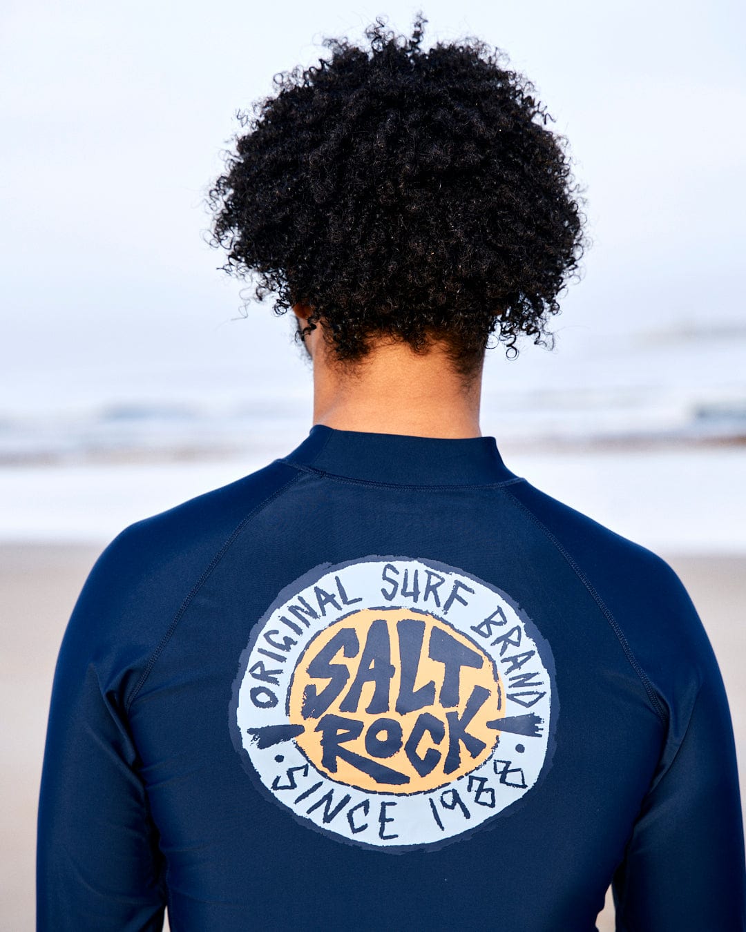 Person wearing SR Original - Recycled Mens Long Sleeve Rashvest - Blue with the Saltrock logo on the back, standing on a beach, facing away from the camera.