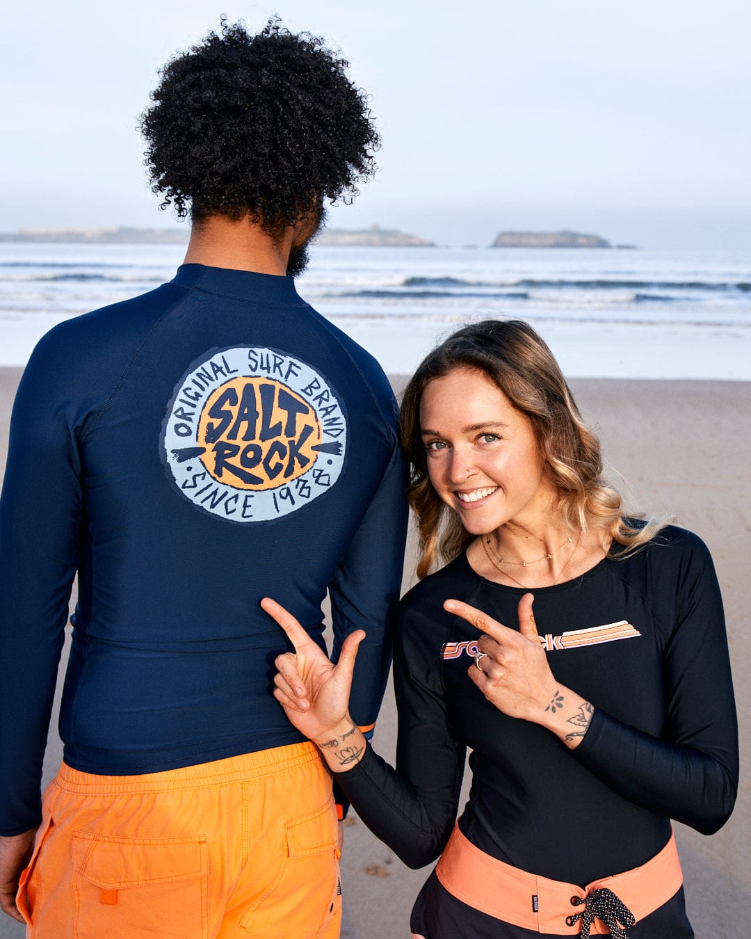 Two people on a beach, one making a shaka sign, wearing surf apparel including boardshorts with a logo "Saltrock" visible on a shirt. They are smiling and seem happy.