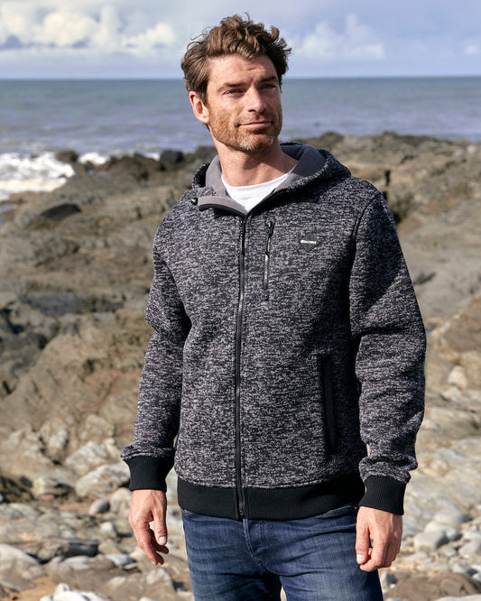 The man is standing on a rocky beach - his outfit includes a stylish and comfortable Saltrock Oskar - Mens Bonded Zip Hoodie - Grey made from textured knit material, perfectly suited for the cool breeze.