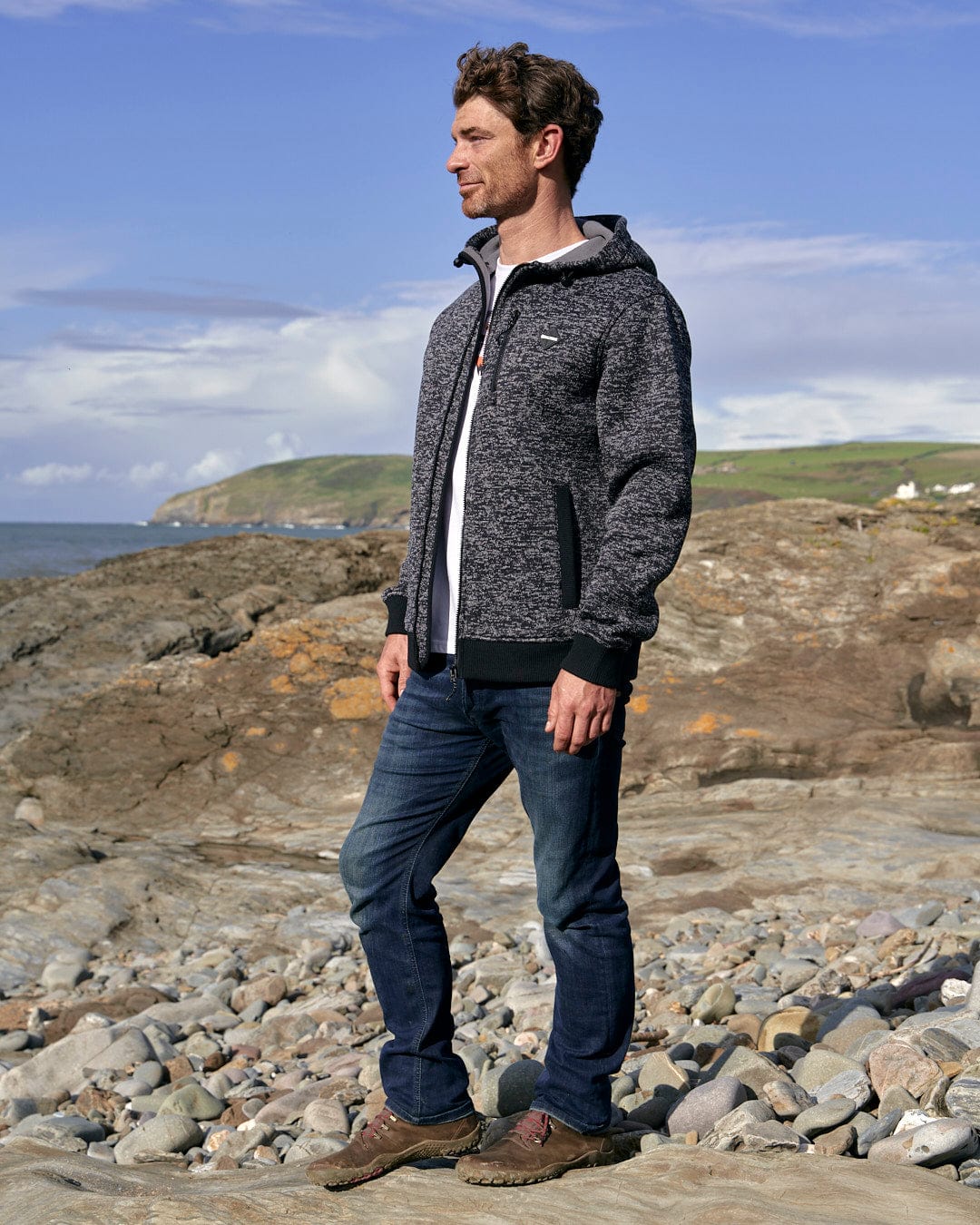 A man wearing a Saltrock Oskar - Mens Bonded Zip Hoodie - Grey standing on a rocky beach.
