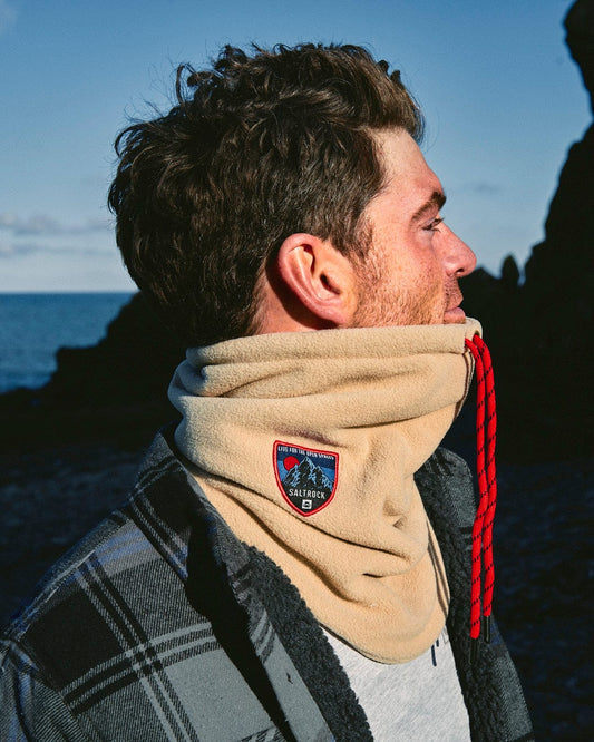 A man wearing a Saltrock Patchy - Borg Snood - Beige near the ocean.