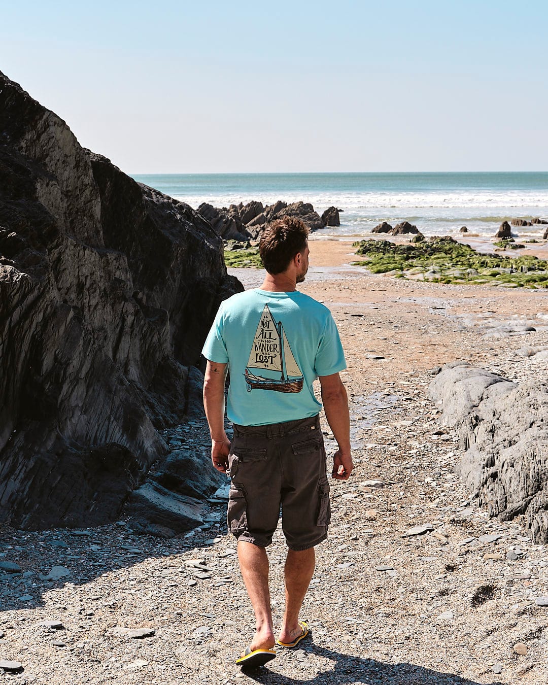 Man standing on a rocky shore, wearing Penwith II - Mens Cargo Shorts in Dark Grey by Saltrock, looking out at the sea.