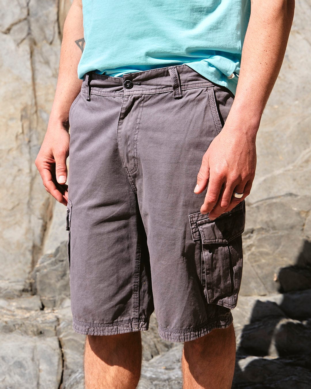 A person standing in Penwith II - Mens Cargo Shorts - Dark Grey by Saltrock with cargo patch pockets against a rocky background.