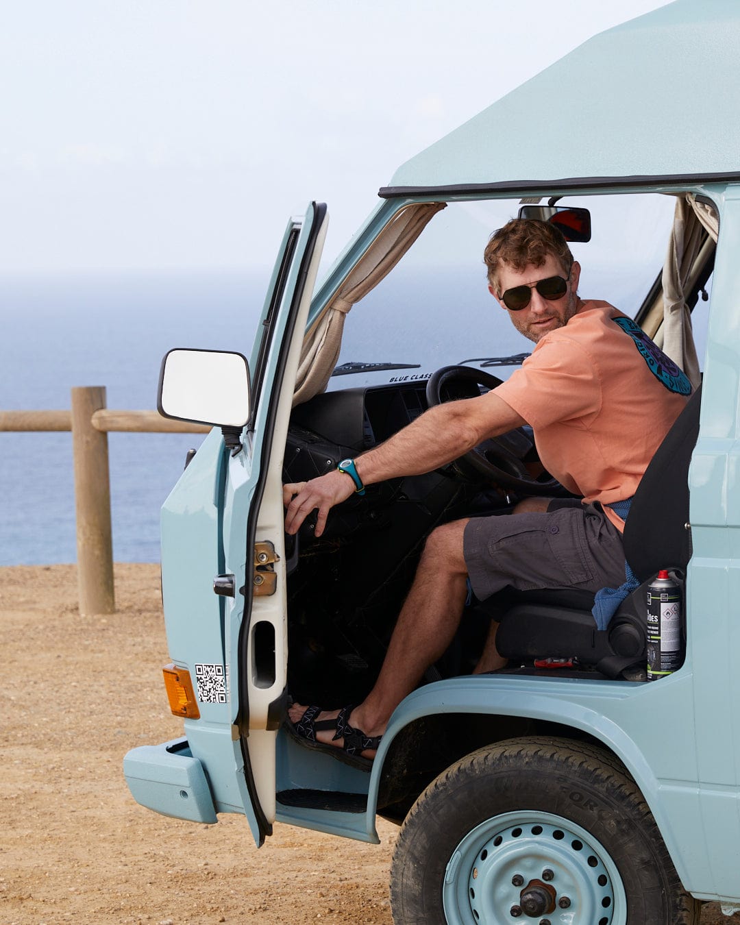A man sitting in the driver's seat of a parked vintage van with the door open, looking back at the camera, wearing Saltrock Penwith II Mens Cargo Shorts in Dark Grey with patch pockets and a unique wash, with a coastal landscape.