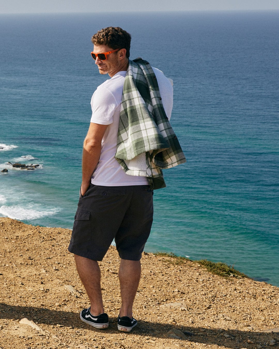 Man with a Penwith II - Mens Cargo Shorts - Dark Grey from Saltrock over his shoulder standing on a coastal cliff overlooking the sea.