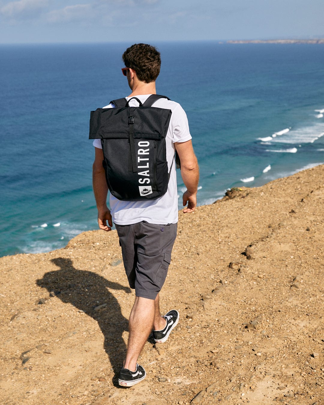 Man with a Saltrock Penwith II - Mens Cargo Shorts - Dark Grey backpack walking along a coastal cliff path.