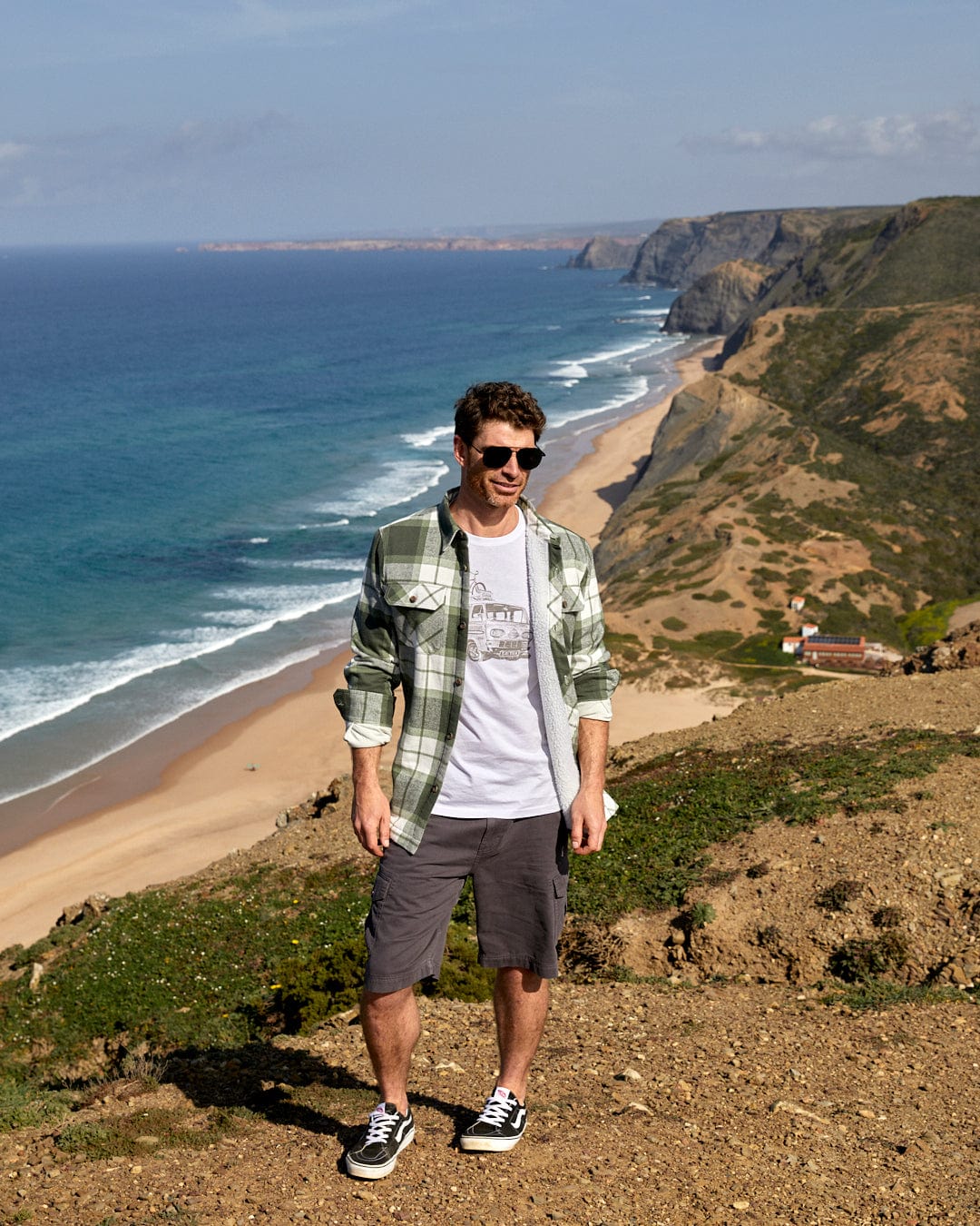 Man standing on a coastal cliff wearing Saltrock's Penwith II - Mens Cargo Shorts - Dark Grey, showcasing scenic ocean and beach views in the background.