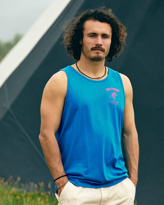 A man with curly hair and a mustache is wearing a blue Pine And Sandy men's vest from Saltrock, made of cotton, standing outdoors in front of a geometric structure.