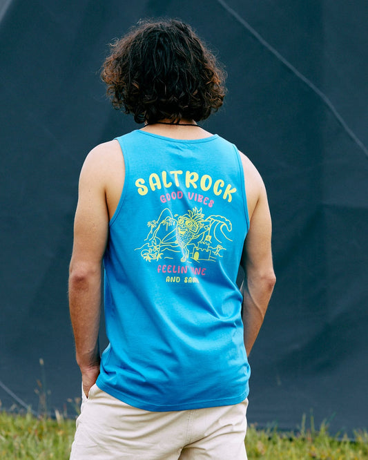 A person with curly hair is wearing the Pine And Sandy Mens Vest in blue by Saltrock, featuring the text "Good Vibes, Feeling Nice and Sunny," paired with beige shorts, standing outdoors against a dark backdrop.