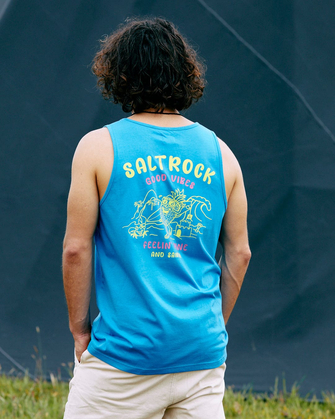A person with curly hair is wearing the Pine And Sandy Mens Vest in blue by Saltrock, featuring the text "Good Vibes, Feeling Nice and Sunny," paired with beige shorts, standing outdoors against a dark backdrop.