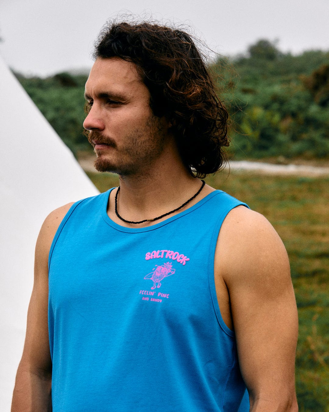 A person with long hair and a slight beard, wearing the Pine And Sandy - Mens Vest - Blue by Saltrock, stands outdoors near greenery, looking to the left.