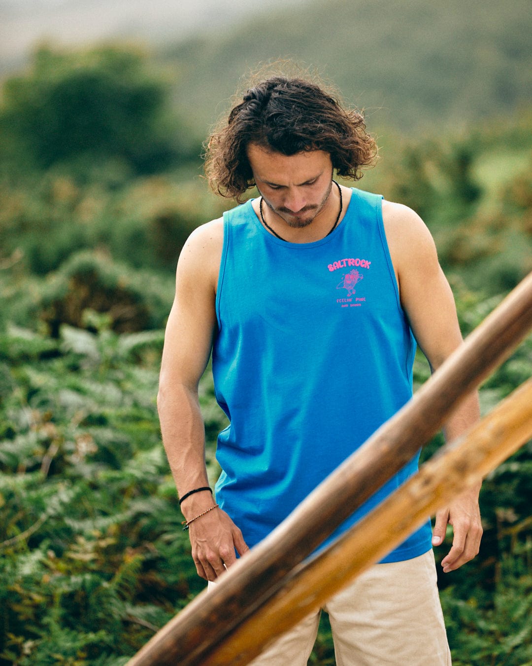 A man with medium-length hair, dressed in the Pine And Sandy men's vest in blue from Saltrock and beige cotton shorts, stands outdoors with greenery in the background. He looks down, and wooden poles are visible in the foreground.