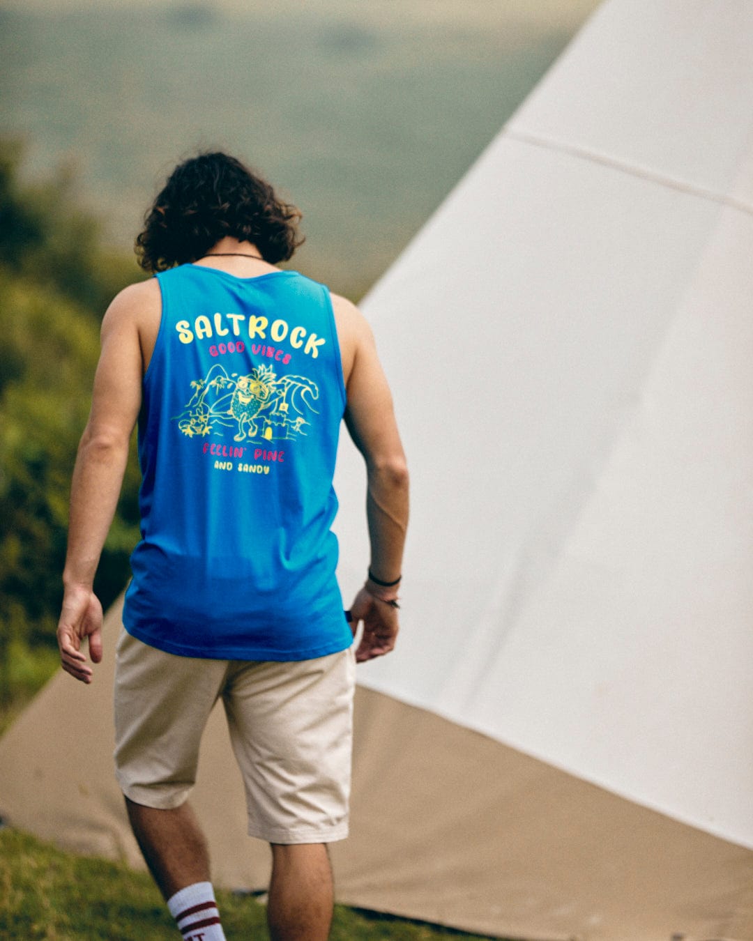 A person with curly hair wearing a blue "Pine And Sandy" tank top by Saltrock and beige shorts stands in front of a beige tent outdoors.