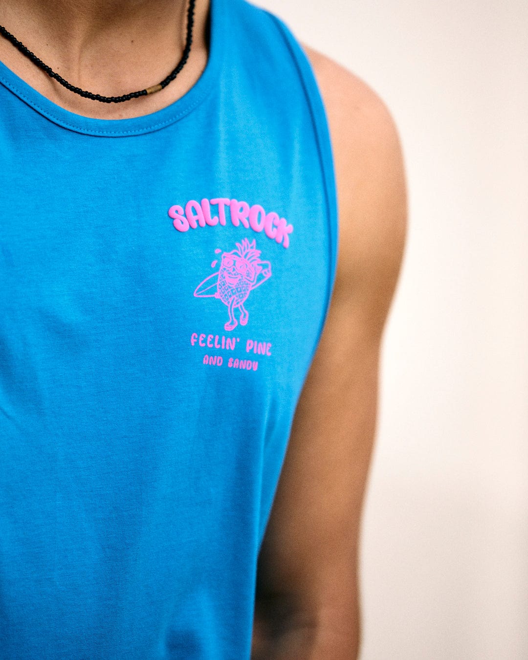 Person wearing the Pine And Sandy - Mens Vest in blue from Saltrock, featuring "Feelin' Pine and Sandy" and "Saltrock" printed in pink on the front, along with a graphic of a pineapple with sunglasses and a surfboard.