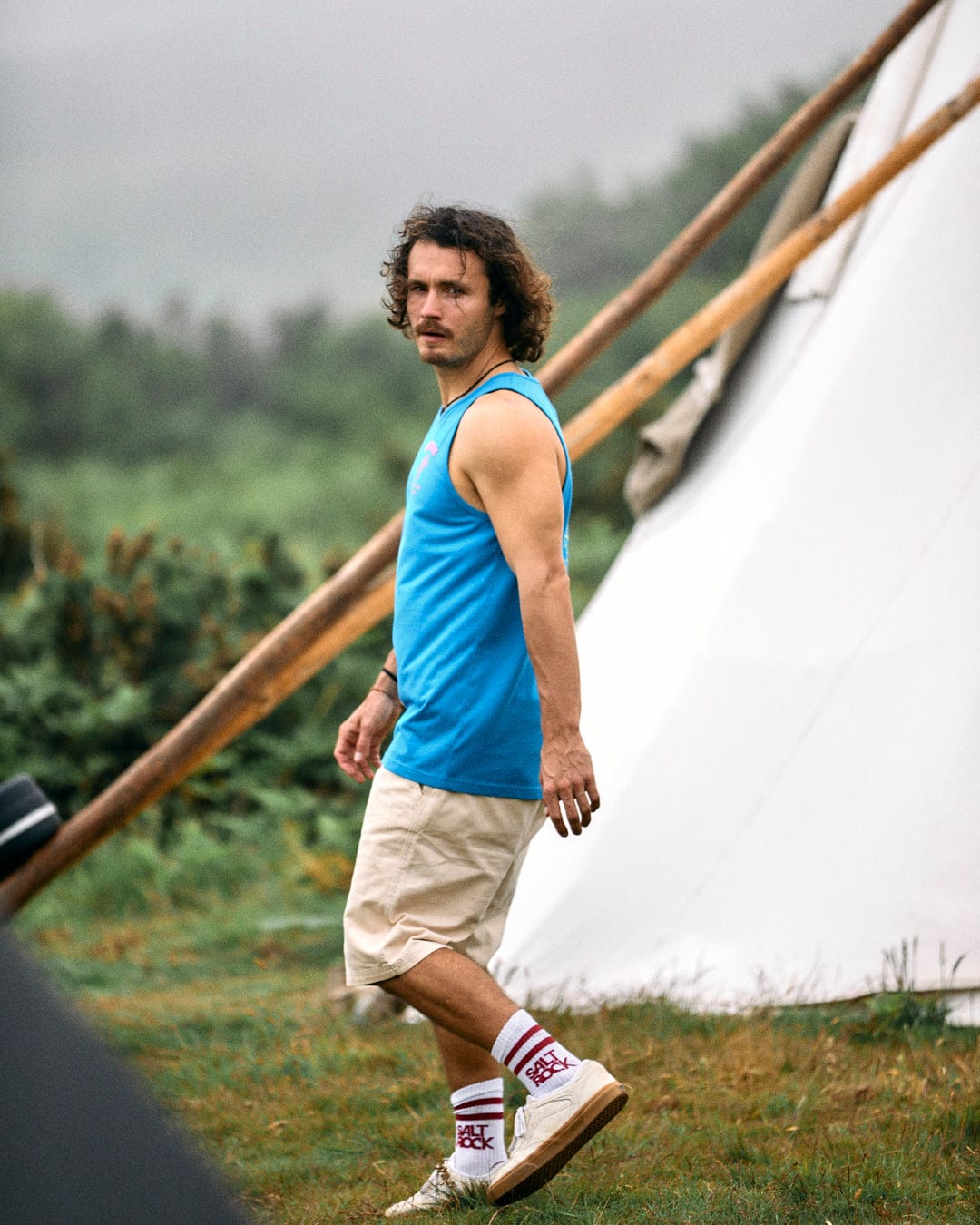 A man with curly hair, wearing a blue Pine And Sandy men's vest from Saltrock made of soft cotton, beige shorts, and white sneakers, stands outdoors near a large white tent on a grassy field.