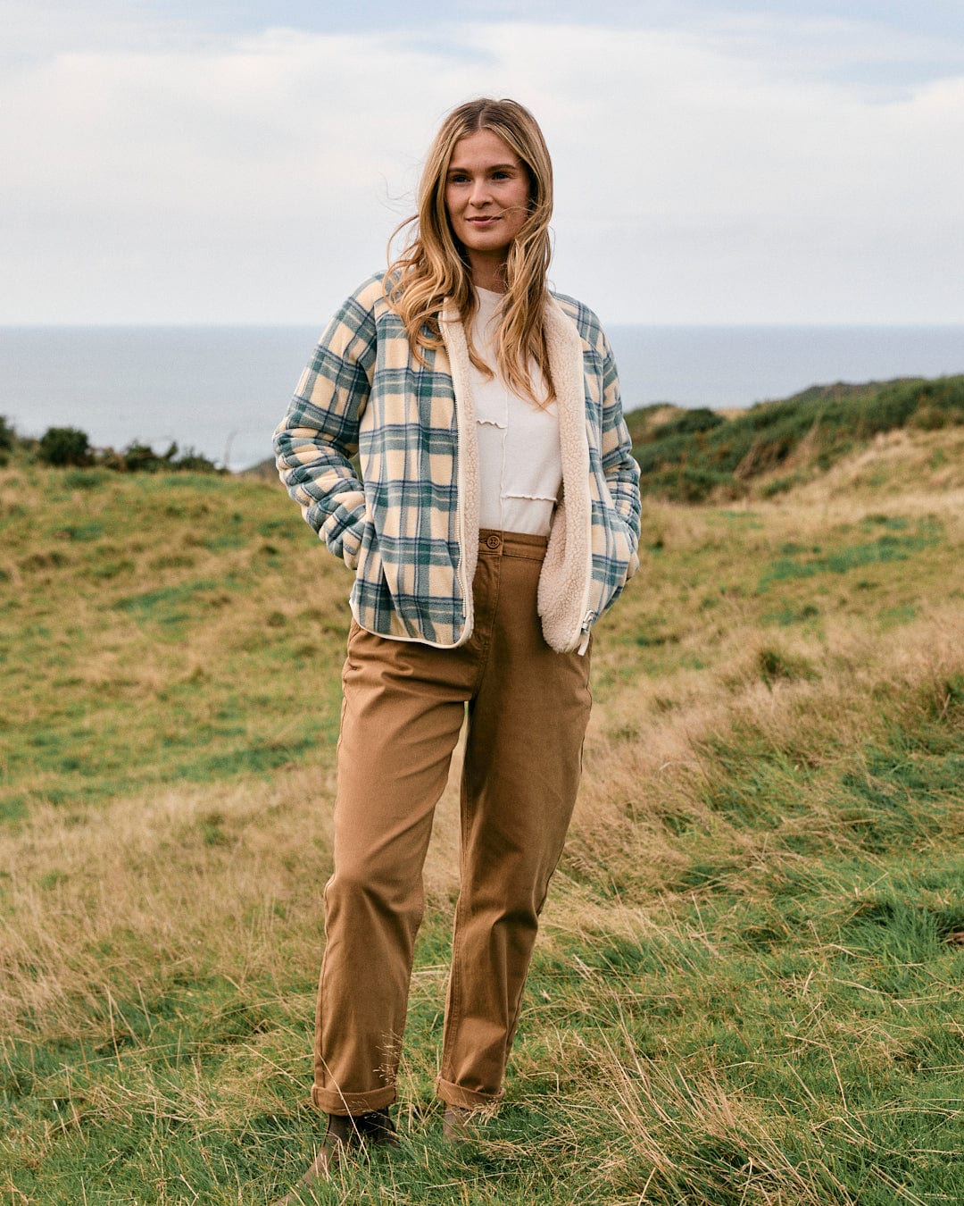 Dressed in the Piper Women's Reversible Zip Collarless Fleece in cream by Saltrock, paired with tan pants and hands in pockets, a person stands on the grassy hillside with the ocean stretching out in the background.