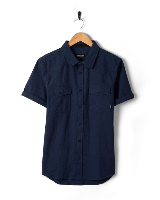 A Polperro - Mens Short Sleeve Shirt - Blue with a Saltrock woven label, hanging on a wooden hanger against a white background.