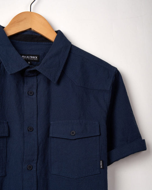 Saltrock Polperro - Mens Short Sleeve Shirt - Blue made from waffle textured fabric on a wooden hanger against a white background, featuring buttoned pockets and a visible Saltrock woven label.