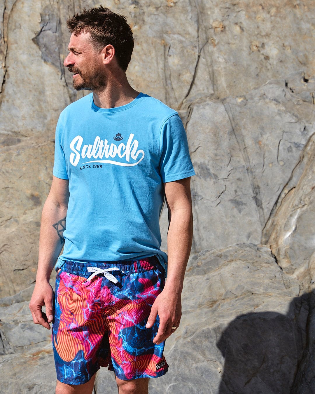 A man wearing a Saltrock Poolside - Mens All Over Print Swimshort - Teal t-shirt and floral shorts.
