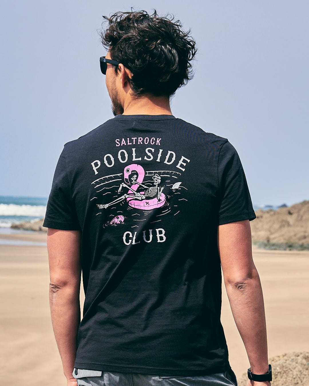 A man standing on the beach wearing a black t-shirt that says Saltrock - Poolside Club  - Mens Short Sleeve T-Shirt - Black.