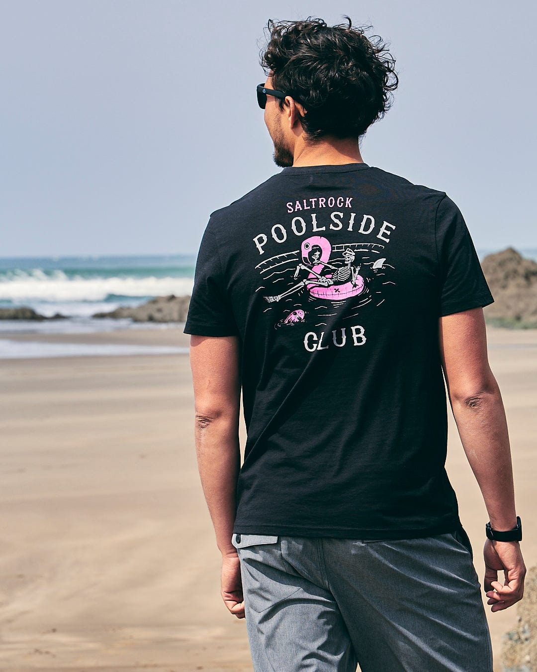 A man walking on the beach wearing a Saltrock Poolside Club Mens Short Sleeve T-Shirt in Black with a pink flamingo on it.