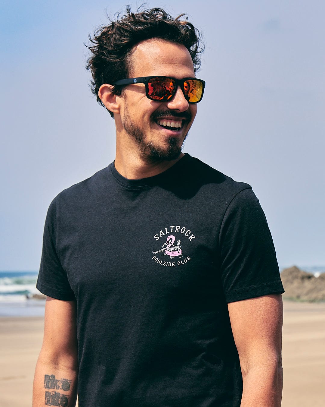 A man wearing sunglasses and a Saltrock Poolside Club Mens Short Sleeve T-Shirt - Black on the beach.