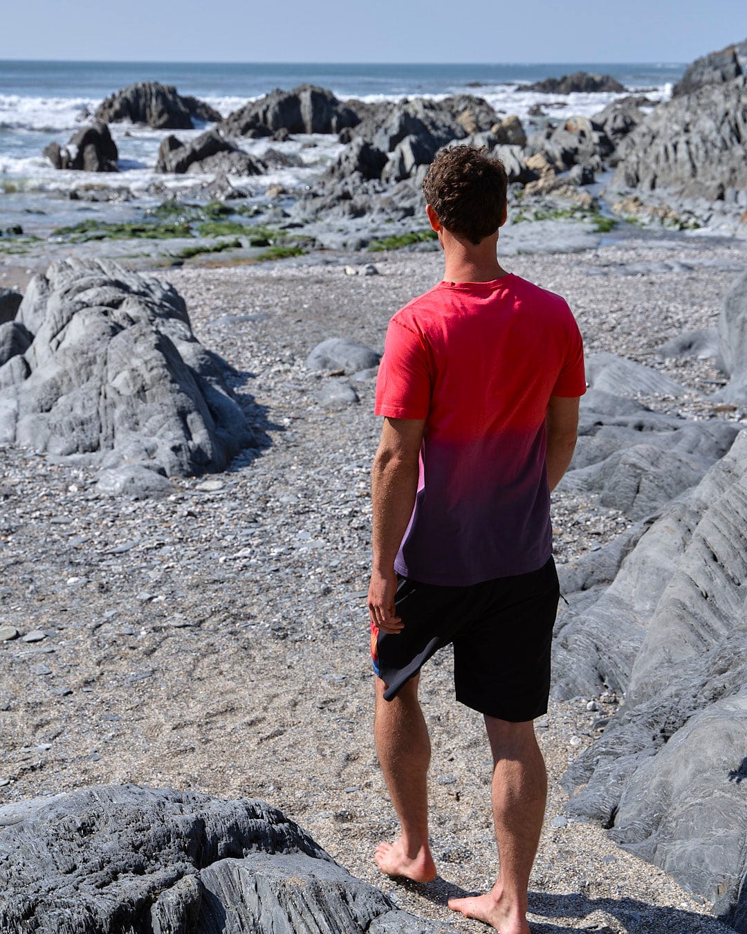 A man wearing a Saltrock Poolside - Mens Dip Die Short Sleeve T-Shirt - Pink, standing on a beach looking at the ocean.
