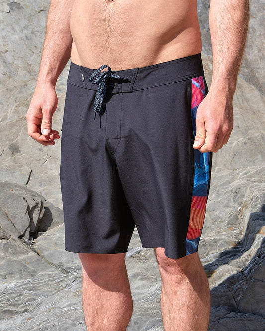 A man wearing Saltrock's Poolside Panel - Mens Recycled 4-Way Stretch Boardshort - Black standing on a rock.