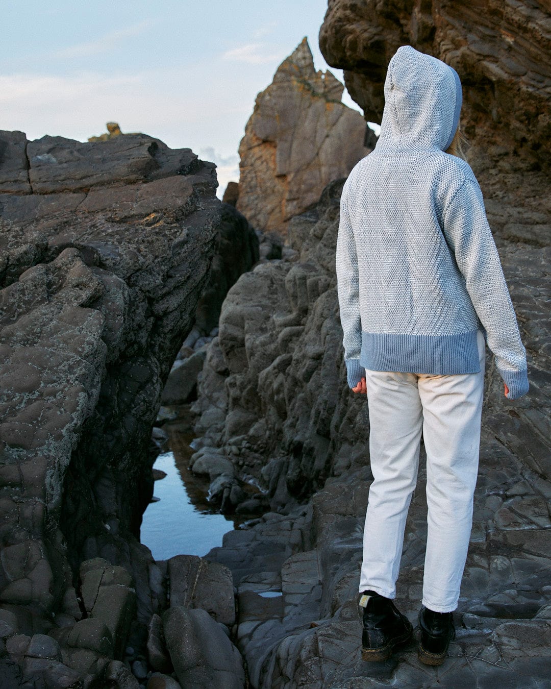 A person wearing the Poppy - Womens Knitted Pop Hoodie - Blue standing on a rocky shore, showcasing Saltrock branding.