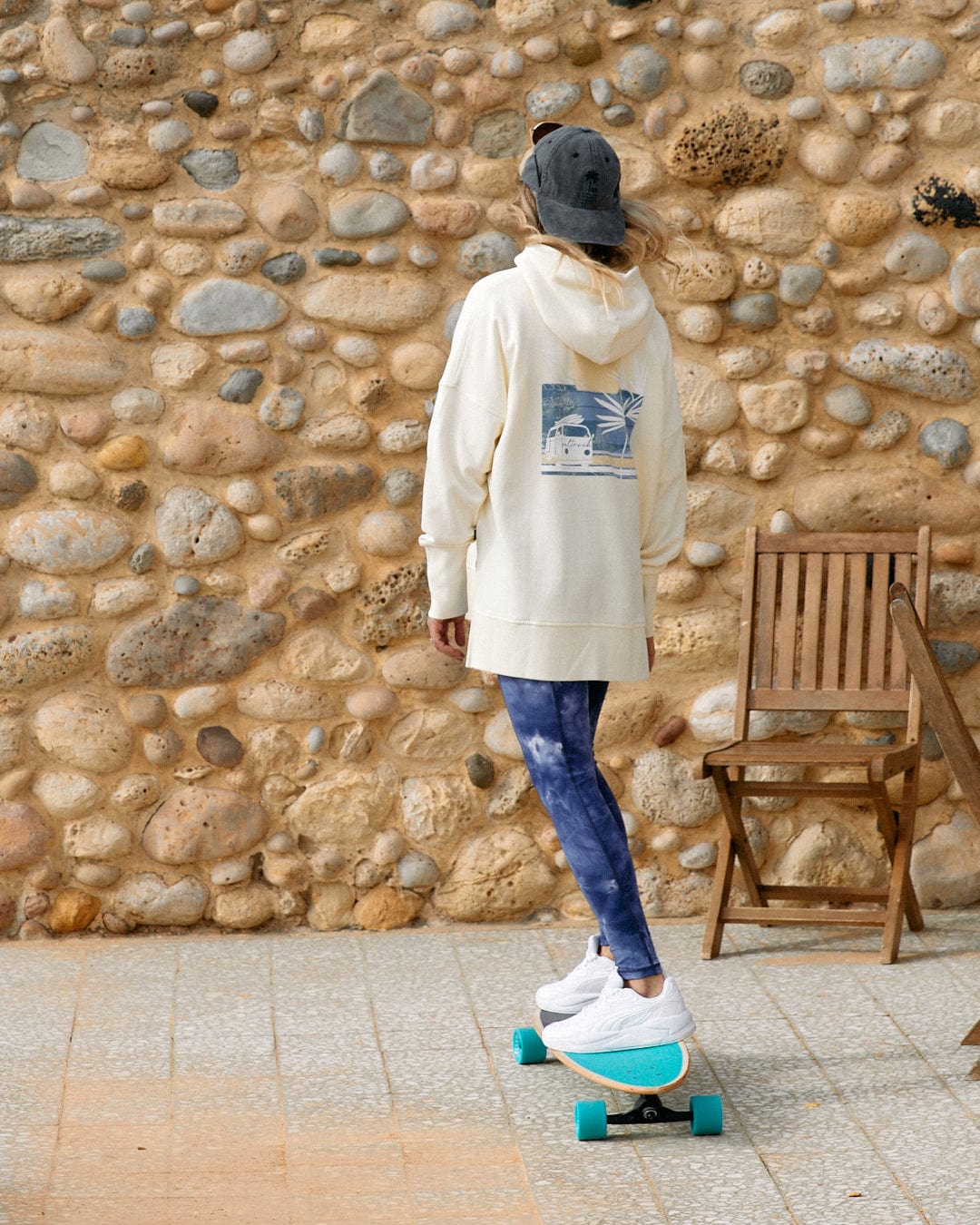A person riding a skateboard near a stone wall and a wooden chair, with Saltrock Poster - Womens Pop Hoodie - Cream branding visible.