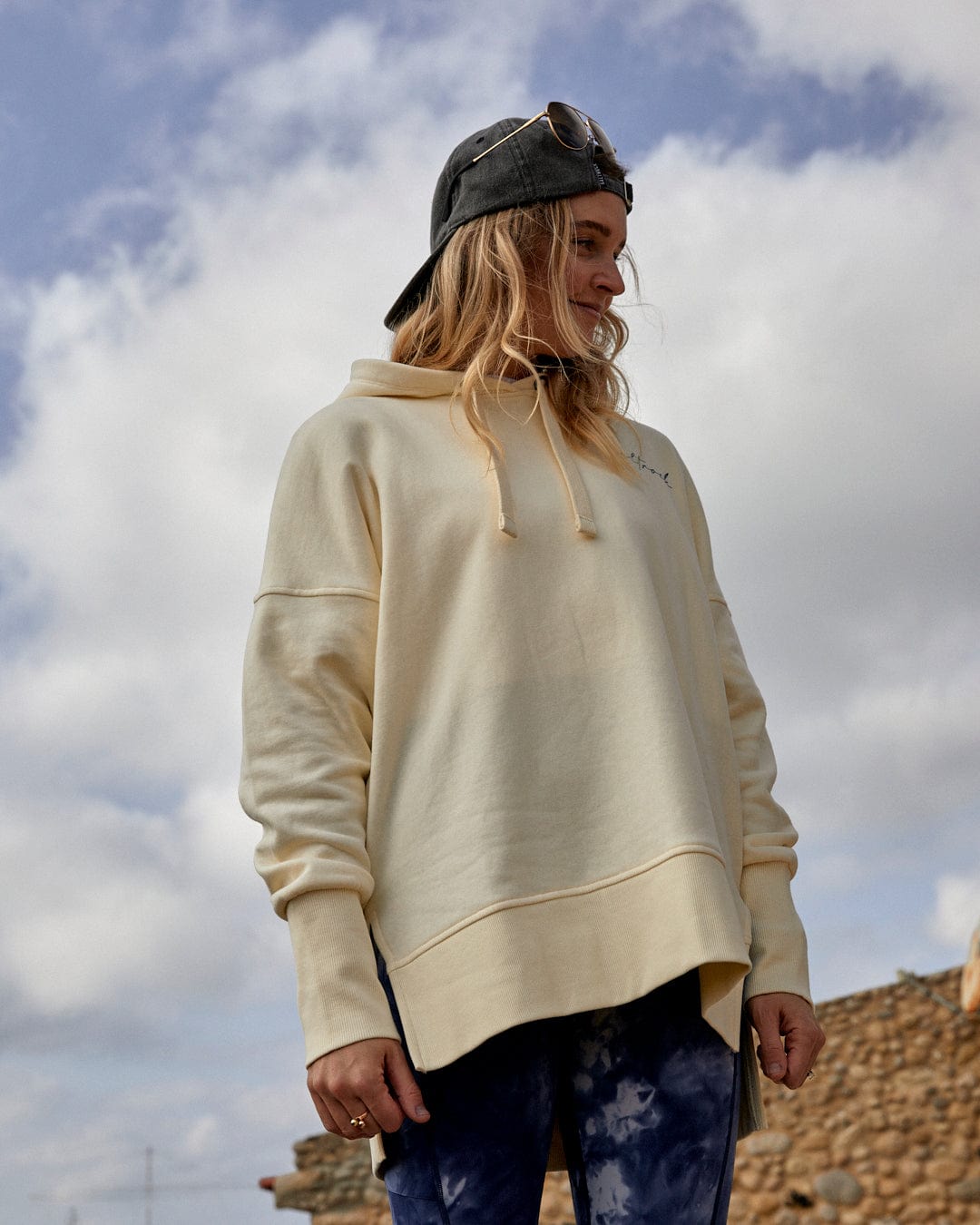 Person in Womens Pop Hoodie in Cream and Saltrock branding cap standing outdoors with cloudy skies in the background.
