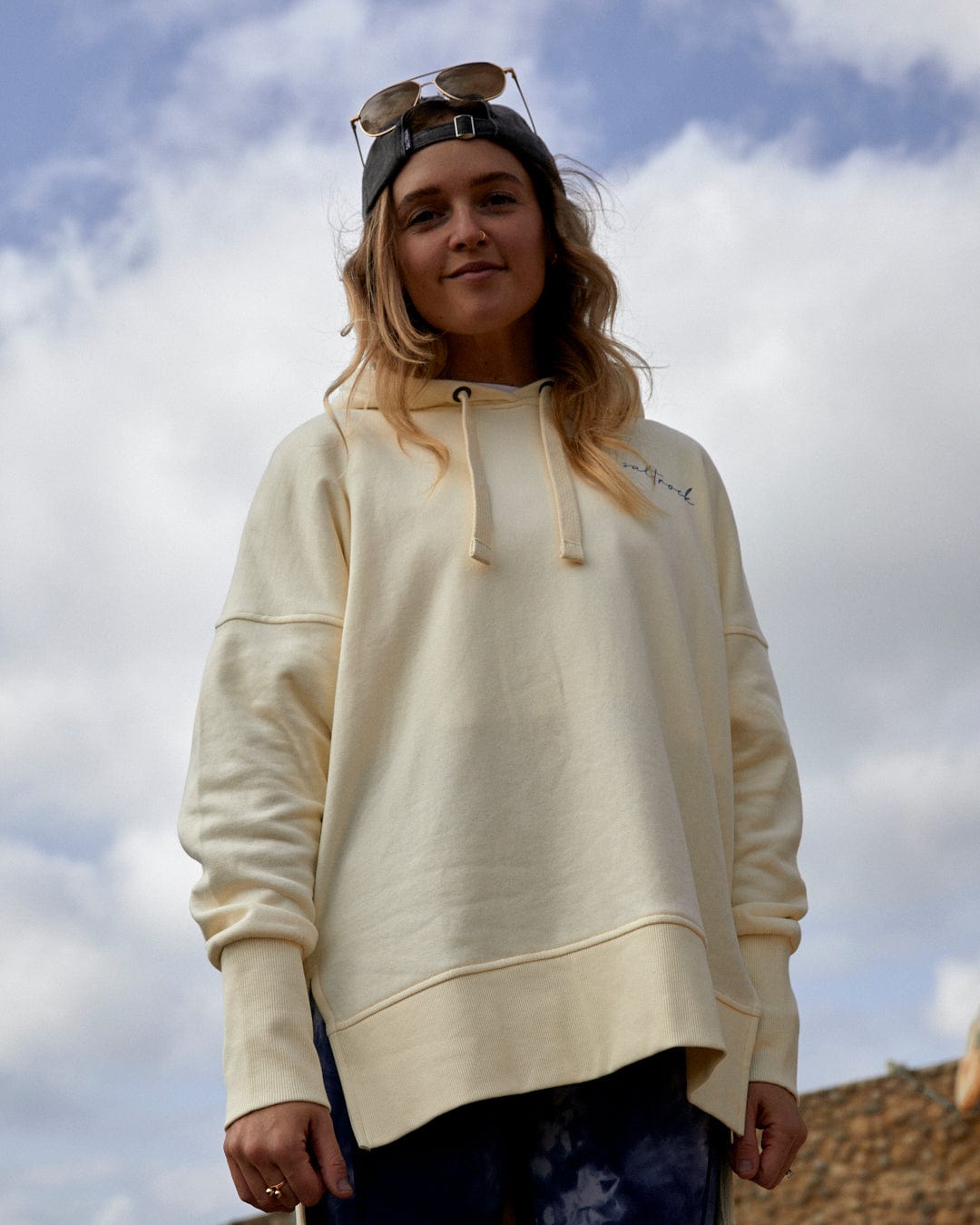 Woman in a Saltrock cream hoodie with Saltrock branding and sunglasses posing against a cloudy sky.
