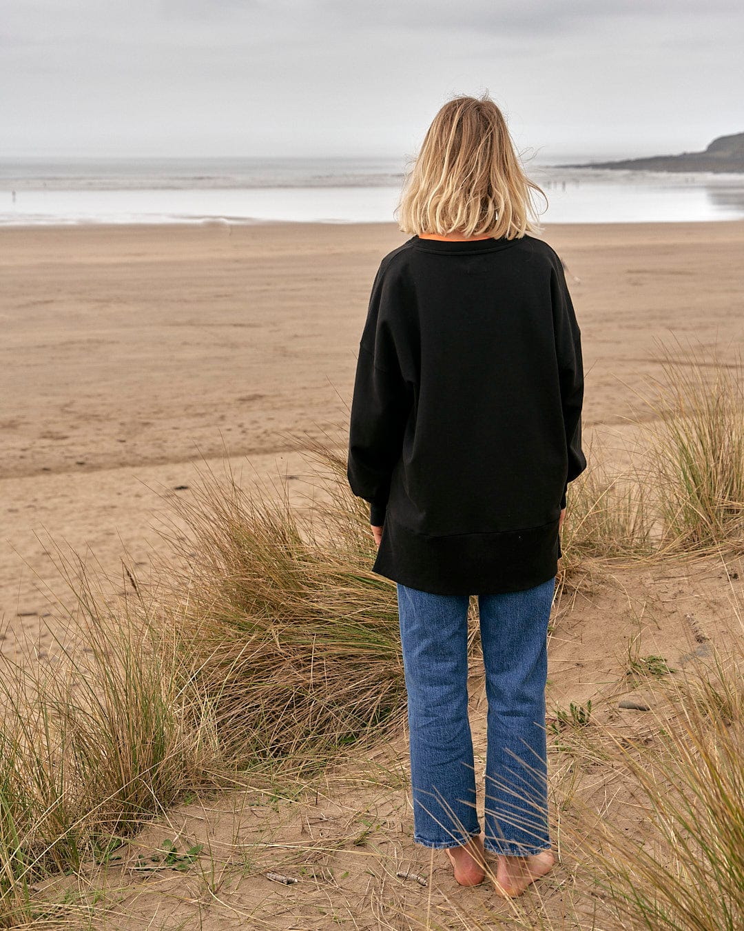 A woman standing on a beach looking at the ocean while wearing the Saltrock Purfect Wave Gradient - Womens Boyfriend Fit Sweat - Black.