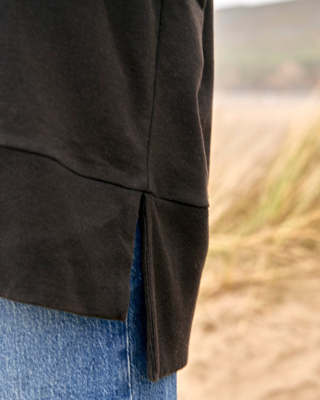 A person wearing Saltrock's Purfect Wave Gradient - Womens Boyfriend Fit Sweat - Black jeans and a black hoodie on a beach.