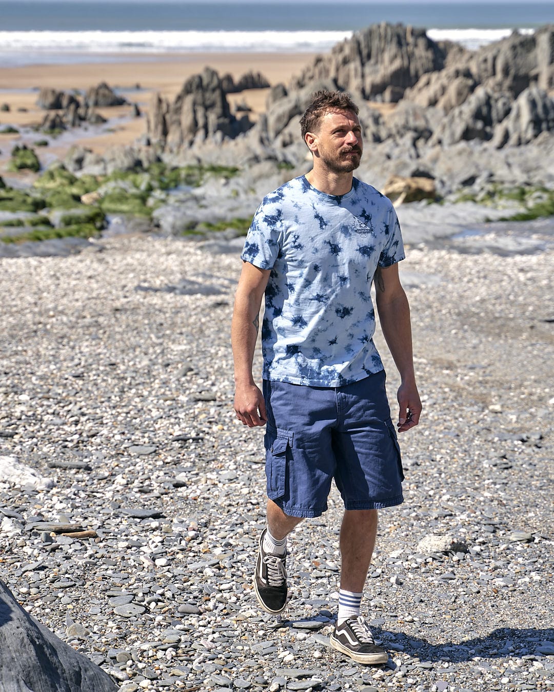 A man walking on the beach with a Saltrock Purfect Wave - Mens Tie Dye Short Sleeve T-shirt - Light Blue skateboard.