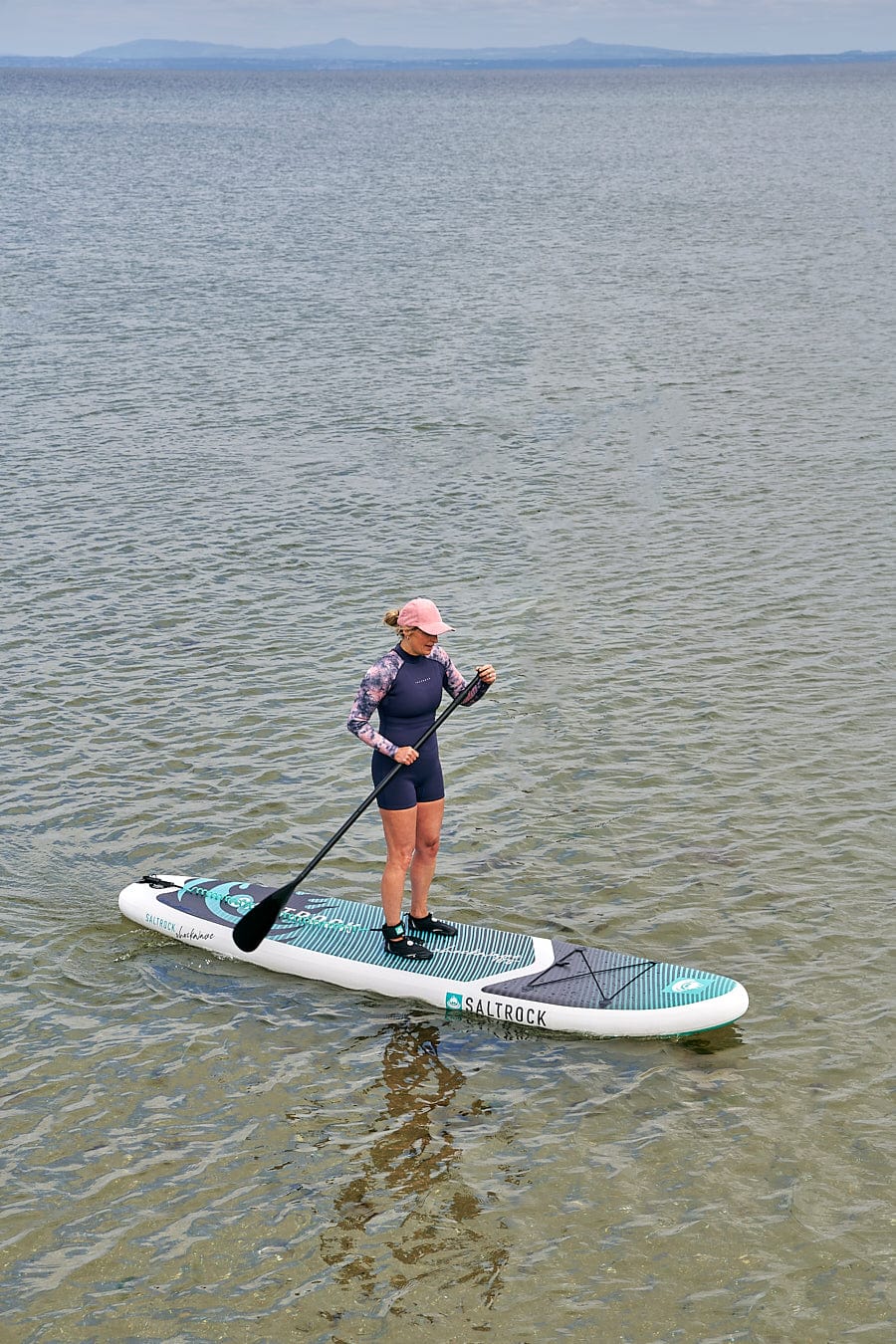 A woman standing on a Saltrock Shockwave - Inflatable Stand Up Paddle Board - Turquoise in the water.